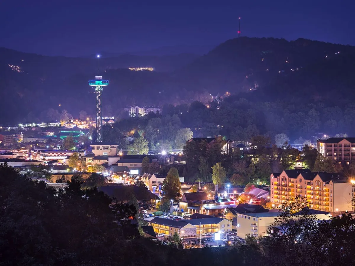 A lively nighttime view of downtown Gatlinburg, featuring warmly illuminated buildings, mountain backdrops, and a tower adorned with lights. The scene captures the town’s vibrant energy and inviting ambiance after dark.