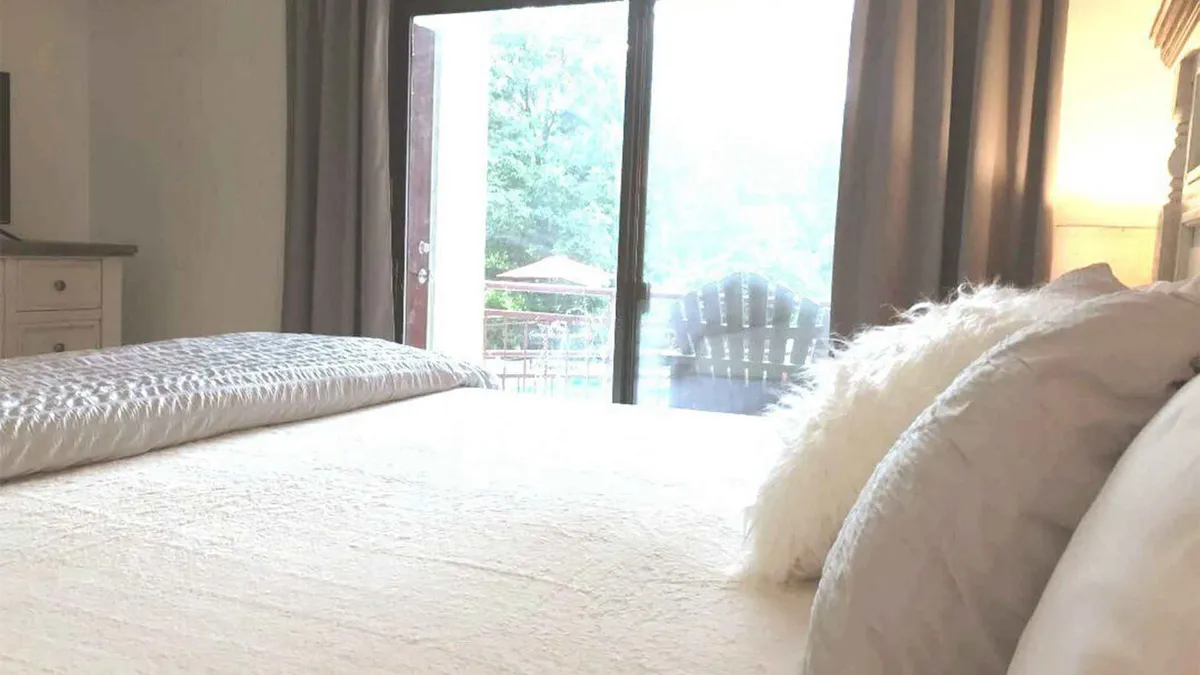 A light-filled bedroom with textured bedding, a large window offering an outdoor view, a soft throw pillow, and cozy accents. The space is styled for comfort, inviting relaxation and quiet moments.