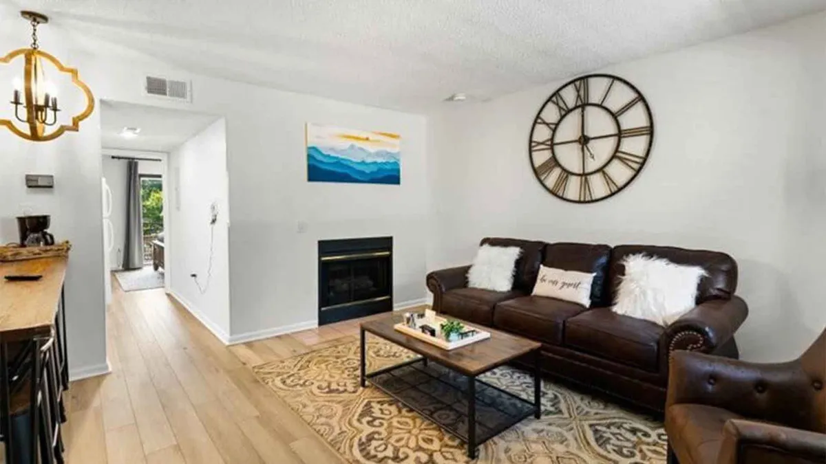 A cozy and stylish living room featuring a leather sofa, coffee table, large wall clock, and curated artwork on the wall. The ambiance is warm and welcoming, with comfortable seating ideal for relaxing or socializing, and the glow of a nearby fireplace adds to the atmosphere.