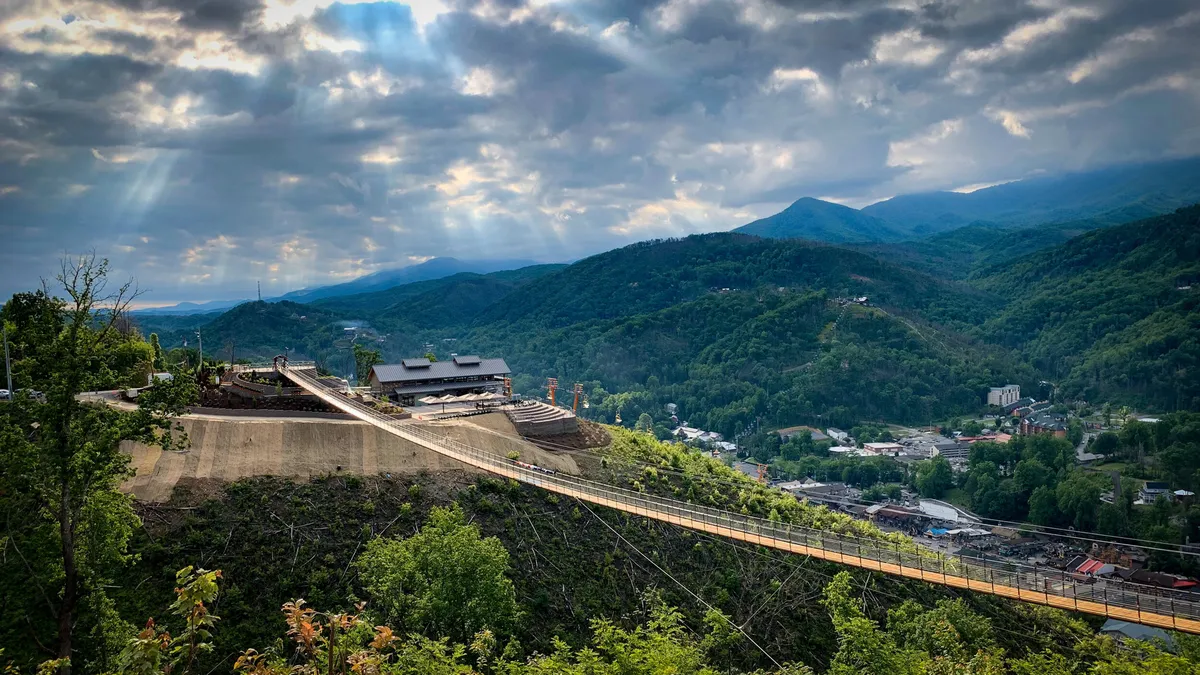 A panoramic mountain landscape featuring a striking suspension bridge, dense forests, and a modern building nestled in the hills. The scene exudes adventure, with the bridge inviting exploration and the surrounding terrain hinting at nearby hiking and zip-lining opportunities.