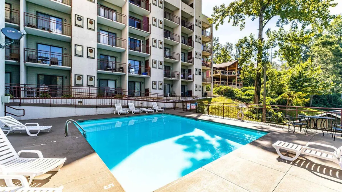 A wide shot of a sparkling swimming pool bordered by lounge chairs and surrounded by lush greenery and balconies. The setting conveys a sense of community and leisure, with inviting spaces for swimming, sunbathing, and socializing.