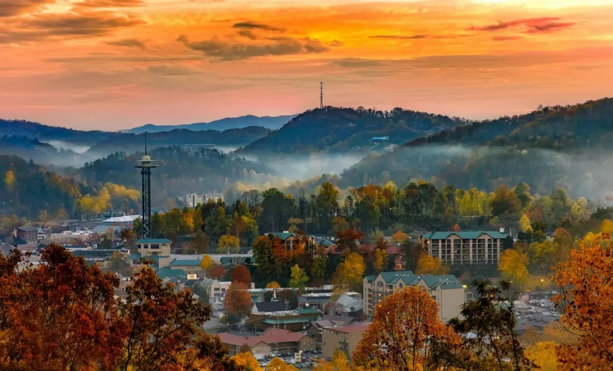 An expansive vista of distant mountains, a quaint town below, and vibrant autumn foliage glowing beneath a sunset sky. An observation tower stands as a focal point, suggesting opportunities for sightseeing and leisurely exploration.