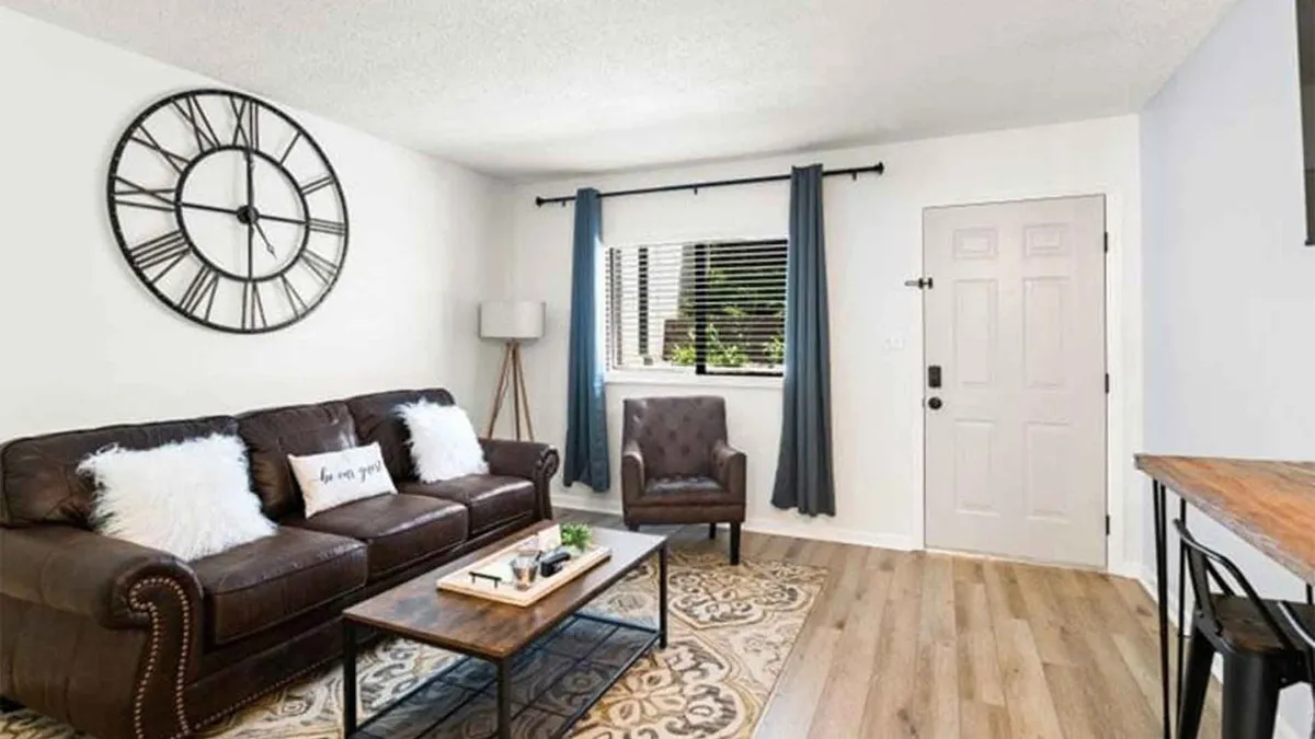 Another perspective on the living area, featuring a large wall clock, a comfortable sofa, coffee table, armchair, and a window with curtains letting in soft natural light. The setup is perfect for hosting guests, reading, or sharing special moments together.