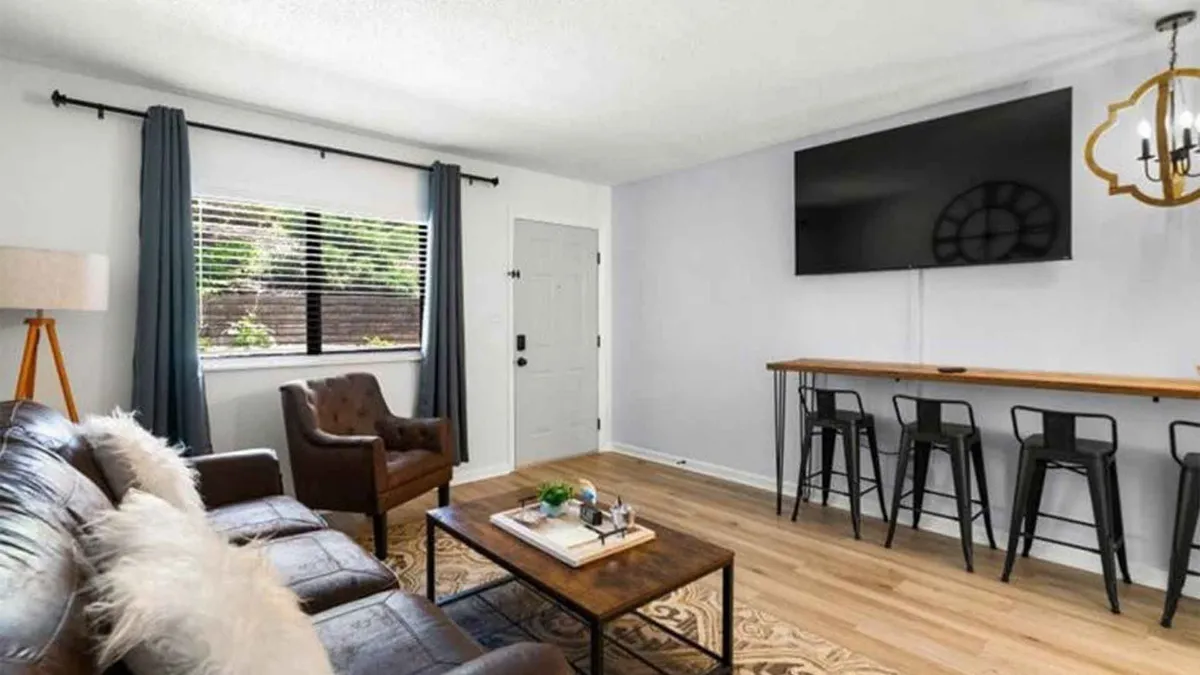 A modern open-plan living and dining area anchored by a leather sofa and wooden coffee table. A flat-screen TV, bar stools at a breakfast bar, and a window with curtains suggest a space designed for both relaxation and socializing. The mood is cozy, inviting, and contemporary.