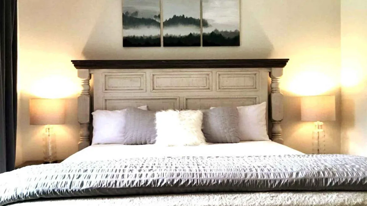 A tranquil bedroom scene featuring a plush bed with a prominent headboard, soft bedding, tasteful artwork above the headboard, and warm table lamps on bedside tables. The mood is serene and calming, suggesting a restful, private retreat.